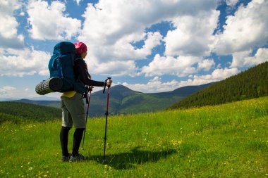 Sırt çantalı ve sopalı kadın gezgin, dağ ve beyaz bulutlu mavi gökyüzünün arka planındaki yeşil çayırda duruyor. Yaz aylarında dağ manzarası. Dışarıda sağlıklı bir yaşam tarzı..