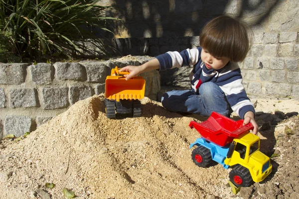 little three year old boy playing in the sand with a digger and dump truck.