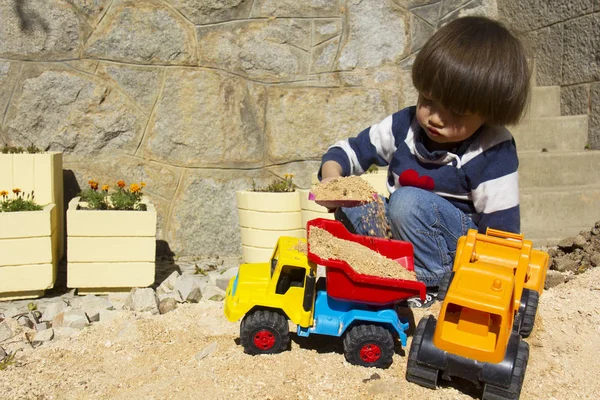 little three year old boy playing in the sand with a digger and dump truck.
