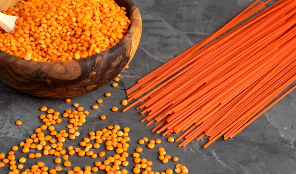 Una ciotola rotonda di legno piena di lenticchie rosse con dentro un cucchiaio di legno. Nelle vicinanze si trovano cereali sparsi e pasta agli spaghetti di lenticchie . — Foto Stock