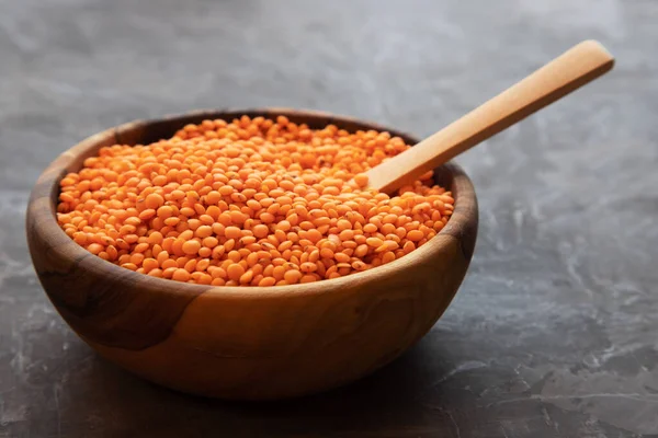 Una ciotola di legno rotonda riempita di lenticchie rosse con un cucchiaio di legno poggia su uno sfondo scuro . — Foto Stock