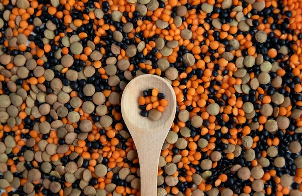 Diverse lenticchie di tre tipi sono sparse sul tavolo e al centro si trova un cucchiaio di legno con fagioli . — Foto Stock