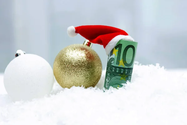 Eine Rolle Banknoten im Wert von 100 Euro in einer Silvesterkappe des Weihnachtsmannes liegen im weißen Schnee neben Weihnachtskugeln in verschiedenen Farben. — Stockfoto