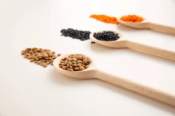 Un cucchiaio di legno con lenticchie marroni primo piano, sullo sfondo ci sono due cucchiai con lenticchie nere e arancioni fuori fuoco. Fondo bianco . — Foto Stock