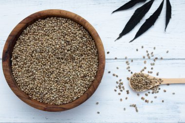 Tahta bir kasede korumasız kenevir tohumları, tohumlu bir kaşık ve beyaz ahşap bir masada bir bitki yaprağı. Süper yemek..