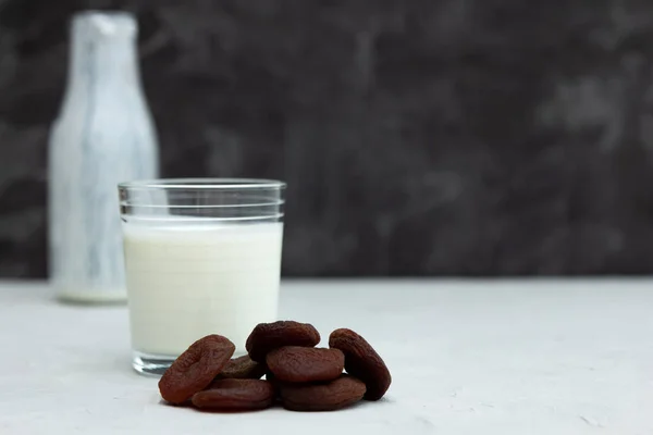 Ein Glas Joghurt, Joghurt oder Ayran auf einem weißen Tisch, daneben liegt eine Handvoll getrocknete Früchte, getrocknete Aprikosen. Im Hintergrund eine Flasche mit einem Getränk. dunkler Hintergrund. gesunde Zwischenmahlzeit — Stockfoto