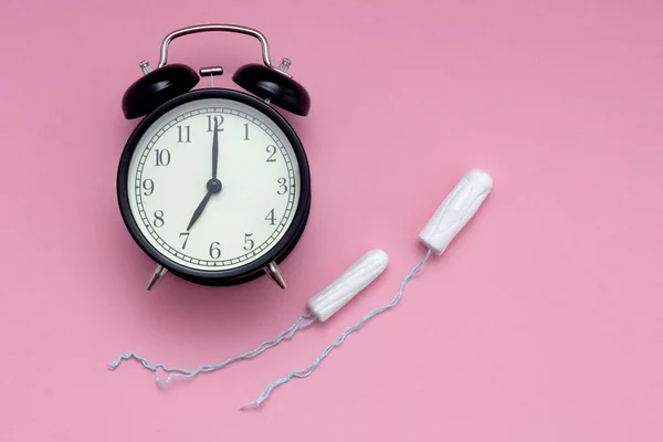 ピンクの背景に黒い目覚まし時計と2つの白いスワブ。女性誌のコンセプト。月経の始まり. — ストック写真