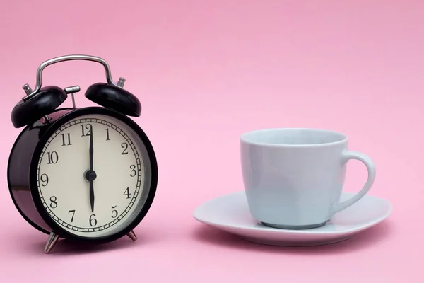 A black alarm clock in retro style and a white coffee cup on a small saucer stand on a pink background. Good morning concept. Morning coffee. — 스톡 사진