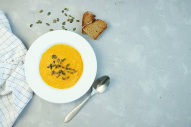 Yanında biberiye ve kabak çekirdekleri olan beyaz bir tabakla balkabağı çorbası. Gri arka planda bir tabak çorba, yanında da bir parça ekmek var. Boşluğu kopyala