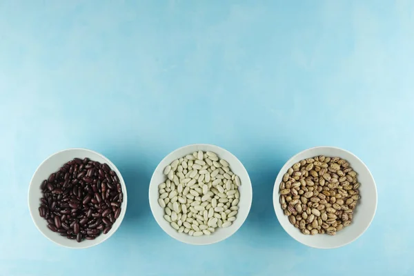 Tres tipos, variedades de frijoles de riñón están en galletas de cerámica blanca, tazas en una mesa azul. Judías coloridas. Lugar para el texto. Fuente de proteína de frijol — Foto de Stock