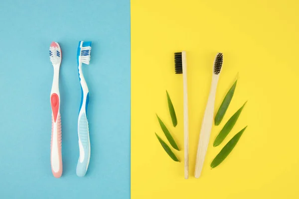 Two plastic toothbrushes and two bamboo toothbrushes with bamboo leaves on a blue and yellow background. Natural materials in everyday life. Oral care — Stock Photo, Image