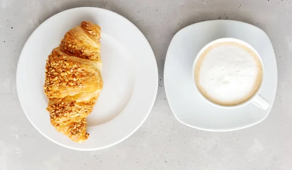 Croissant francés clásico fresco, espolvoreado con nueces, sobre un plato de porcelana blanca y una taza blanca de capuchino, café con espuma al lado. Fondo gris. Vista desde arriba. Desayuno fresco . — Foto de Stock