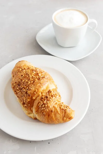 Croissant francés fresco en un plato blanco y una taza de café blanco con espuma cerca. Desayuno francés clásico. Postre de Almuerzo. Orientación vertical — Foto de Stock