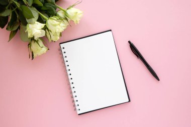 Not ve çizimler için bir defter pembe arka planda, siyah kalem ve açık yeşil güllerin yanında. Bir mektup, bir mesaj, bir not