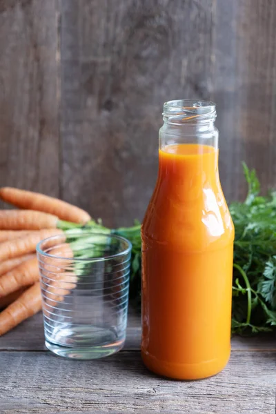 Succo di carota in una bottiglia di vetro e un bicchiere vuoto nelle vicinanze. Sullo sfondo un mazzo di carote fresche con cime verdi. Fondo in legno. Orientamento verticale — Foto Stock