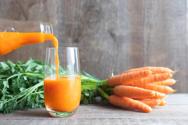 El jugo de zanahoria se vierte de una botella de vidrio en un vaso, un montón de zanahorias frescas con tapas verdes se encuentra en la parte posterior. Nutrición dietética. Estilo de vida saludable. Fondo de madera —  Fotos de Stock