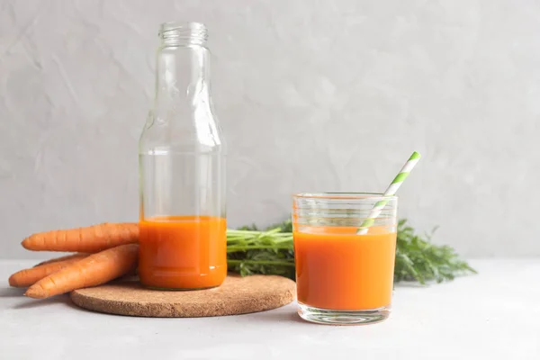Een glazen fles met vers wortelsap op een kurkstandaard en een glas met sap en een cocktailrietje in de buurt. Op de achtergrond een stel verse wortelen. Plaats voor tekst. — Stockfoto