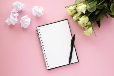 Temiz beyaz çarşaflı bir defter, üzerinde siyah bir kalem, pembe bir arka plan ve buruşuk beyaz çarşaflar ve yakınlarda bir buket gül var. Kayıt, rapor, mesaj, mektup. Metin için yer