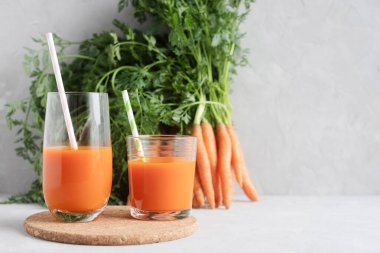 İki bardak dolusu havuç suyu ve kokteyl tüpleri mantar tezgahında duruyor. Arka planda üst kısımları taze havuç dolu. Metin için yer.