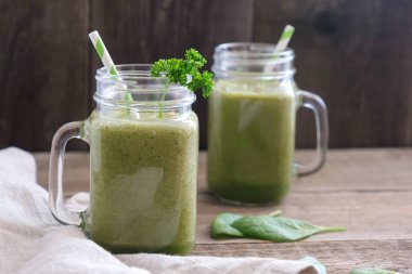 İki bardak, bardak, yeşil smoothie ve kokteyl tüpleri ahşap bir masada duruyor. Keten peçete ve ıspanak yapraklarının yanında..