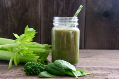 Yeşil smoothie ve bambu kokteyli pipetli bir bardak ahşap bir tahtanın üzerinde duruyor. Yakınlarda kereviz, ıspanak ve maydanoz yaprakları var. Faydalı bir atıştırma. Vegan, vejetaryen içeceği. Boşluğu kopyala.