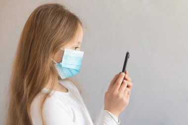 Tıbbi maske takan kahverengi saçlı şirin küçük bir kız elinde telefonla ayakta duruyor. Kendini izole etme ve virüs salgını sırasında iletişim kurma. Boşluğu kopyala.