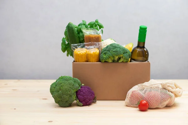 Cardboard box with products, wine, pasta, basil, lemons and fruit products nearby in reusable environmental shopping bags. Home delivery concept. Place for text
