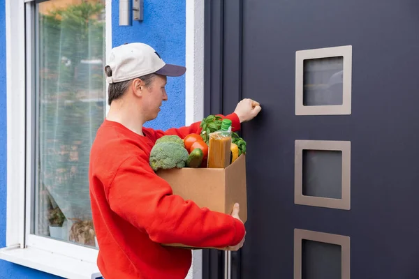 Un livreur de nourriture ou un bénévole portant un pull rouge et un bonnet frappe à la porte avec une boîte de produits. Concept d'aide avec nourriture, livraison à domicile de nourriture pendant la quarantaine . — Photo