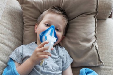 Kahverengi saçlı küçük üzgün bir çocuk kanepede yatarken solunum yapıyor. Ev tedavisi.