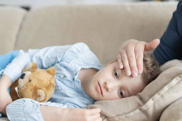 A sick and sad cute girl lies on a sofa with a soft toy and her mother checks the heat or temperature on her forehead. sick children, flu — Stock Photo, Image