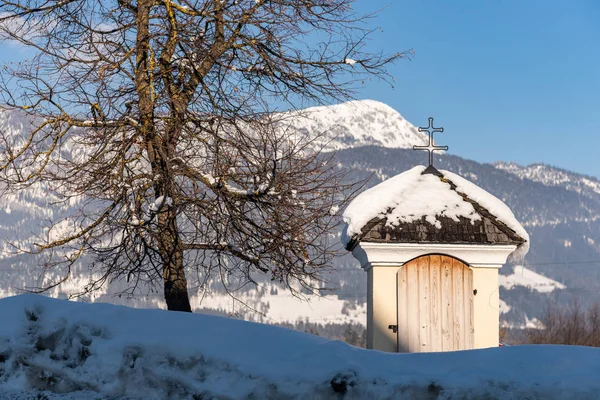 Маленькая деревянная альпийская часовня, покрытая снегом и освещенная теплым солнечным светом. Рядом с ним одинокое дерево. Альпы Австрии — стоковое фото