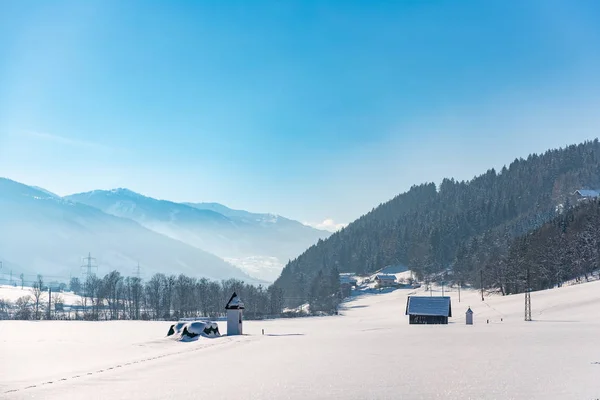 Arkasında ağaçlar ve dağlar olan karla kaplı bir kışın manzarası. Mavi gökyüzü ve küçük bir şapele giden yol. Schladming, Avusturya, Avrupa