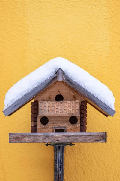 Alimentador casero de madera para pájaros en invierno, bajo la nieve sobre el fondo amarillo de la pared — Foto de Stock