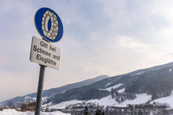 Цепной дорожный знак: Bei Schnee und Eisgltte. Синий предупреждающий сигнал: цепь противоскольжения обязательна. Зимнее время, заснеженная дорога, деревья. Штирия, Австрия — стоковое фото