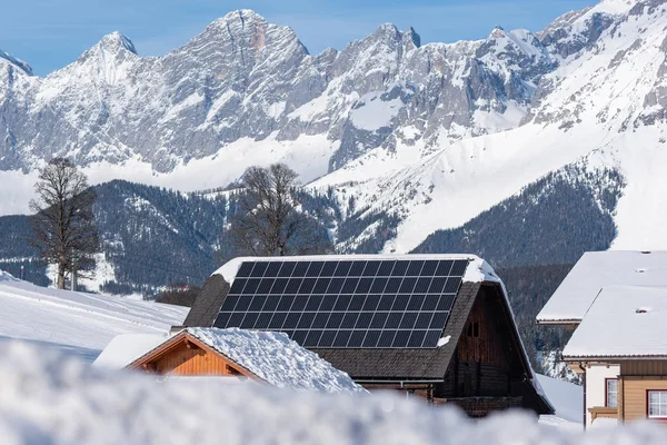 Pannelli fotovoltaici solari fotovoltaici su un tetto di una casa innevata. Elettricità dal sole durante l'inverno. Casa in montagna con montagna innevata sullo sfondo . — Foto Stock