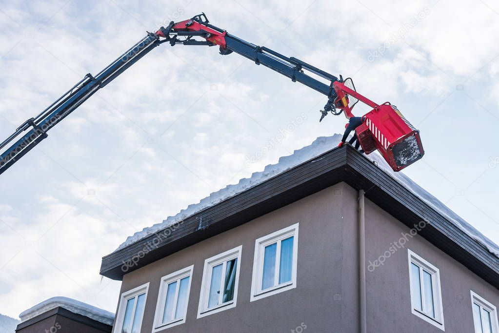 Man in the telescopic aerial platform cleaning snow and ice from roof top, gutter of the building to avoid the danger off falling snow on the pavement, during the winter time.