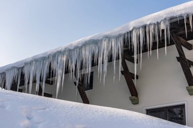 Kışın dağlarda geleneksel ahşap bir evin çatısında yağmur oluğunun üzerinde asılı duran buz saçakları ve kar tehlikeli olabilir. Arka planda mavi gökyüzü.