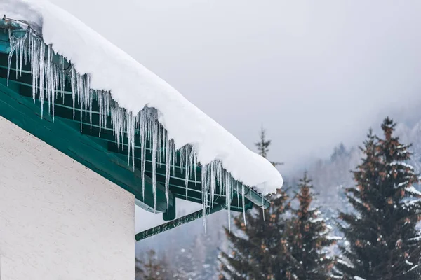 Big icicles and snow hanging over the rain gutter on a roof of a traditional wooden house in the mountains in winter could be dangerous. Trees at the background. Stock Photo
