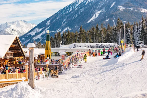 Restaurante alpino lleno de esquiadores, snowboarders en un día soleado. En el fondo estación de esquí de góndola Hochwurzen, montañas nevadas. Austria —  Fotos de Stock