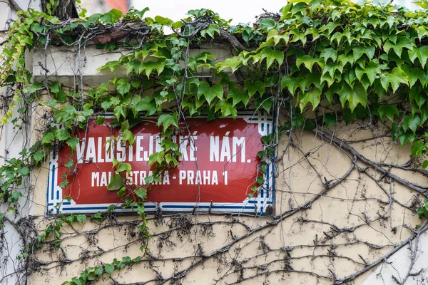 Red street sign on the wall in Prague with white text: Valdstejnske nam, Mala Strana, Prague 1, The Senate of the Parliament of the Czech Republic is located on this street. Ivy growing on the wall.