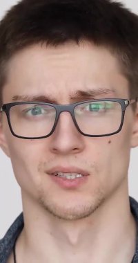 Beyaz arka planda kameraya yüzünü buruşturan gözlüklü bir adamın portresi.