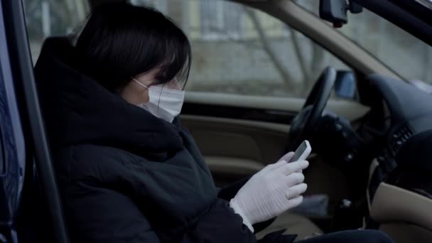 Uma mulher morena de casaco preto está sentada em um carro. Uma mulher está usando uma máscara protetora e luvas. Ela está usando um smartphone . — Vídeo de Stock