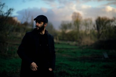 Koyu saçlı, sakallı, doğada sigara içen bir adamın sanatsal portresi. Koyu renk elbiseler, kafasında bir şapka. Film için film işleme.