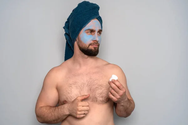 A bearded man with a blue towel on his head and a cosmetic mask on his face. Cosmetology procedures for men. Skin care for men.