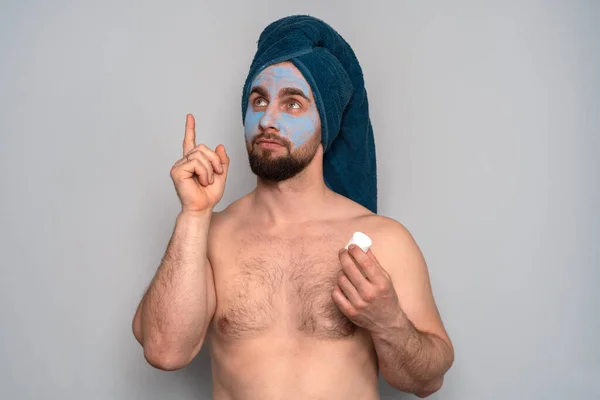 Kafasında mavi havlu ve yüzünde kozmetik maske olan sakallı bir adam. Erkekler için kozmetoloji prosedürleri. Erkekler için cilt bakımı.