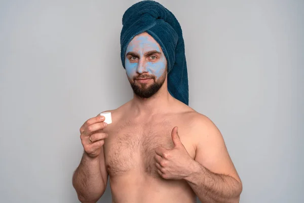 一个留着胡子 头上戴着蓝色毛巾 脸上戴着化妆品的男人 男人的美容程序 男子的皮肤护理 图库图片