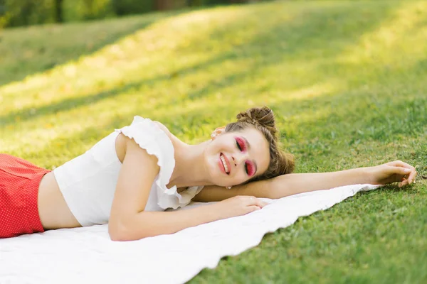 A young girl with bright makeup is lying on a spruce blanket on the grass with her eyes closed and smiling. Professional summer makeup in watermelon style