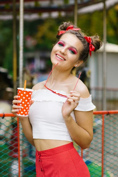 A young girl with bright makeup and a cheerful hairstyle two gulki holds a bright paper Cup in her hands, and in the other hand holds red glasses and . Professional summer makeup in watermelon style