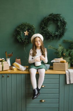 Güzel bir kız elinde beyaz bir sürahi tutuyor ve yeni yıl ve Noel için süslenmiş şık bir mutfakta oturuyor.