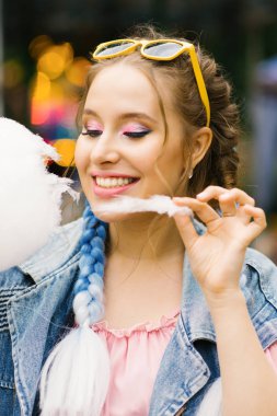 Parlak profesyonel makyajlı, kafasında sarı gözlüklü pamuk helva yiyip gülümseyen bir kız..